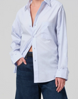 A person is wearing the kayla shirt from Citizens Of Humanity/AGOLDE in light blue, paired with dark blue jeans. The shirt is partially unbuttoned, and the sleeves are slightly rolled up. The person stands against a plain, light gray background.