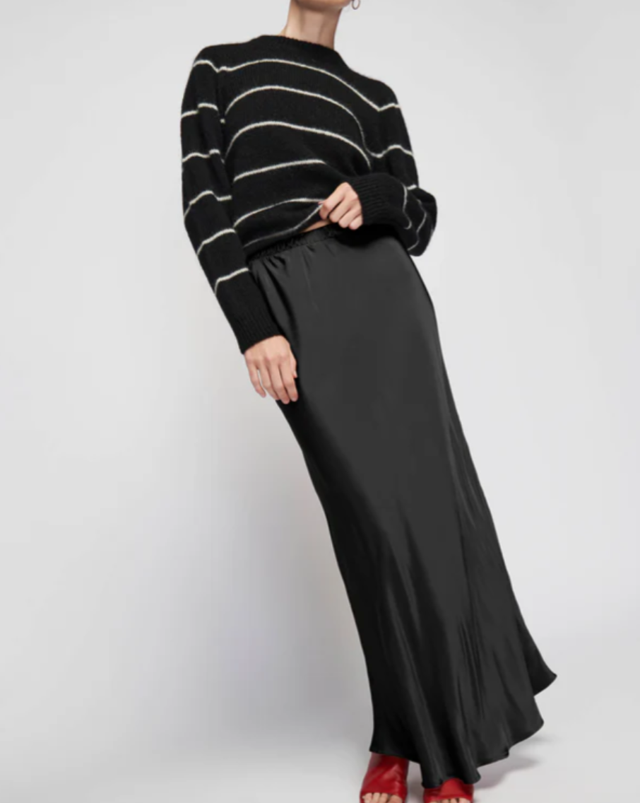 A person is wearing a black and white striped sweater paired with the Gaia Bias Cut Maxi skirt from Nation LTD, which features a long, flowing design and an elastic waist. They are also sporting red shoes against a plain, light-colored background.