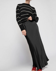 A person is wearing a black and white striped sweater paired with the Gaia Bias Cut Maxi skirt from Nation LTD, which features a long, flowing design and an elastic waist. They are also sporting red shoes against a plain, light-colored background.