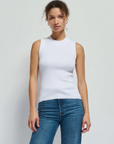 A person with curly hair stands against a plain white background, wearing the Joan Sleeveless Mock Neck top by Nation LTD and blue denim jeans. They have a neutral expression and one arm resting by their side.