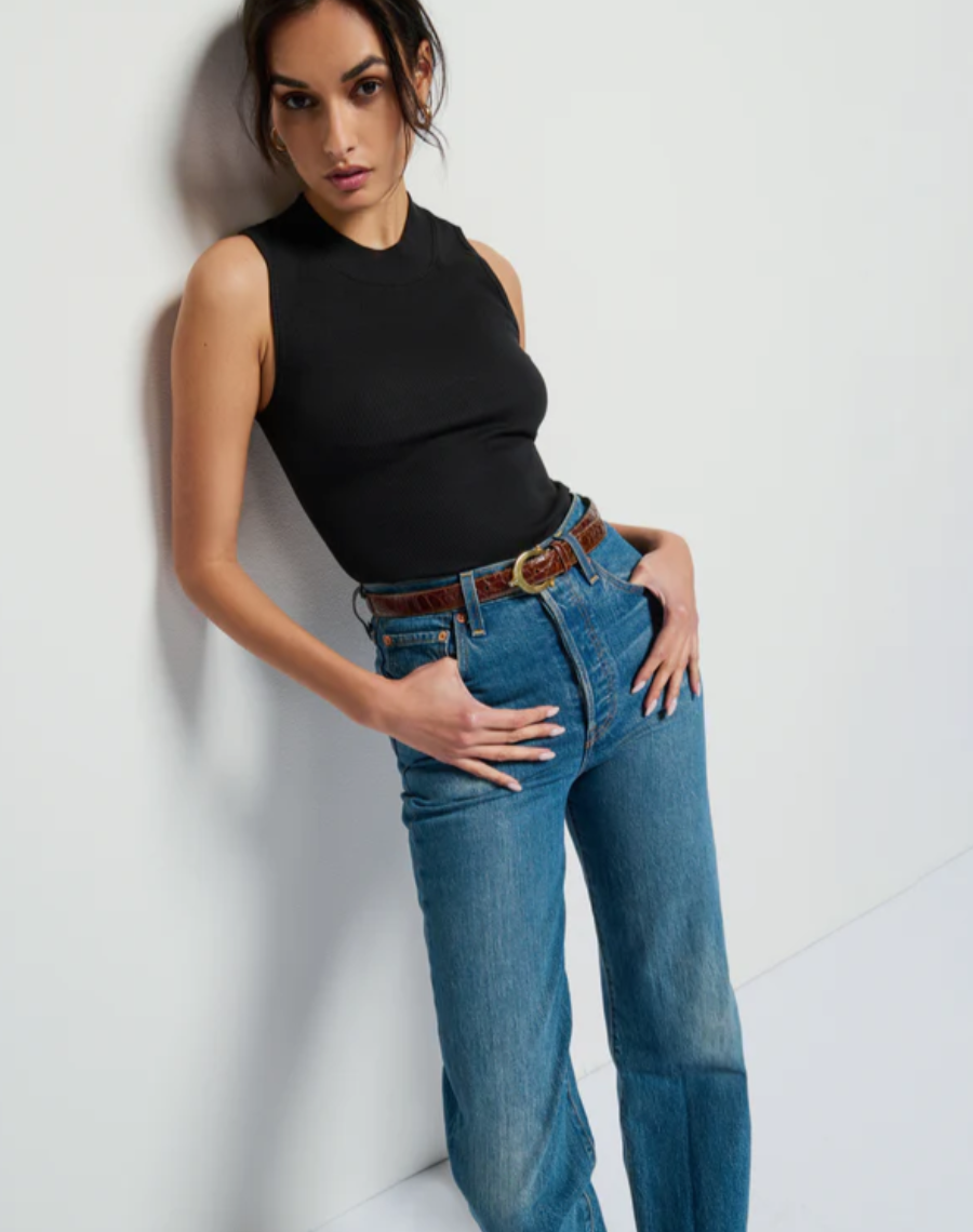 A woman stands against a plain white wall, wearing the Joan Sleeveless Mock Neck by Nation LTD and high-waisted blue jeans. She has her hands in her pockets and is gazing towards the camera. Her dark hair is tied back in a low bun, and she is accessorized with a brown belt.