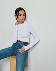 A woman with jet black hair pulled back is wearing the "Gina Slim Mock Neck" white long-sleeve top by Nation LTD, paired with high-waisted blue jeans and a brown belt. She is seated on a wooden block, looking off to the side with one hand touching her neck and the other resting on her leg against a plain white background.