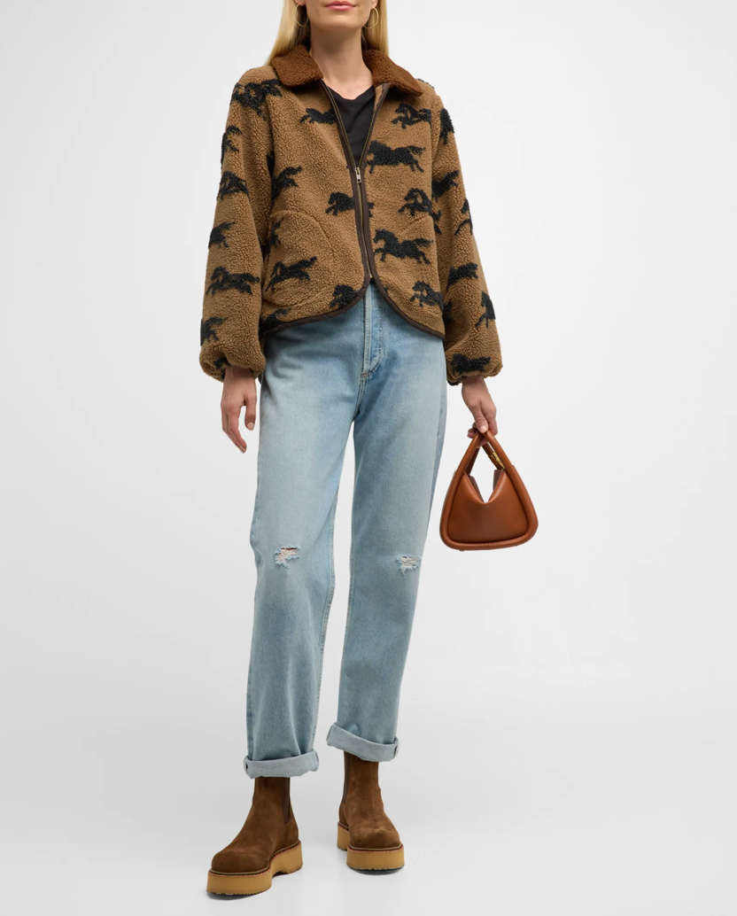 A person wearing "The Pasture Jacket" from The Great Inc., featuring a relaxed fit and horse motif print, paired with a black shirt, light blue distressed jeans, and brown suede boots. They are holding a small, tan geometric-shaped handbag against a plain white background.