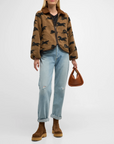 A person wearing "The Pasture Jacket" from The Great Inc., featuring a relaxed fit and horse motif print, paired with a black shirt, light blue distressed jeans, and brown suede boots. They are holding a small, tan geometric-shaped handbag against a plain white background.