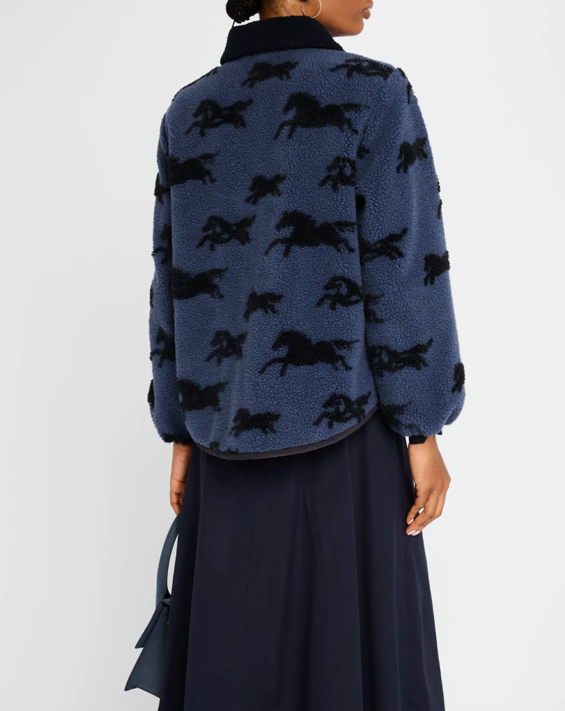 A person wearing The Pasture Jacket by The Great Inc. with a black horse motif and a black skirt. They are holding a navy blue handbag. The view is from the back, showcasing the detailed horse pattern on the relaxed fit jacket.