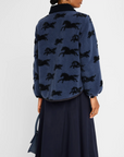 A person wearing The Pasture Jacket by The Great Inc. with a black horse motif and a black skirt. They are holding a navy blue handbag. The view is from the back, showcasing the detailed horse pattern on the relaxed fit jacket.