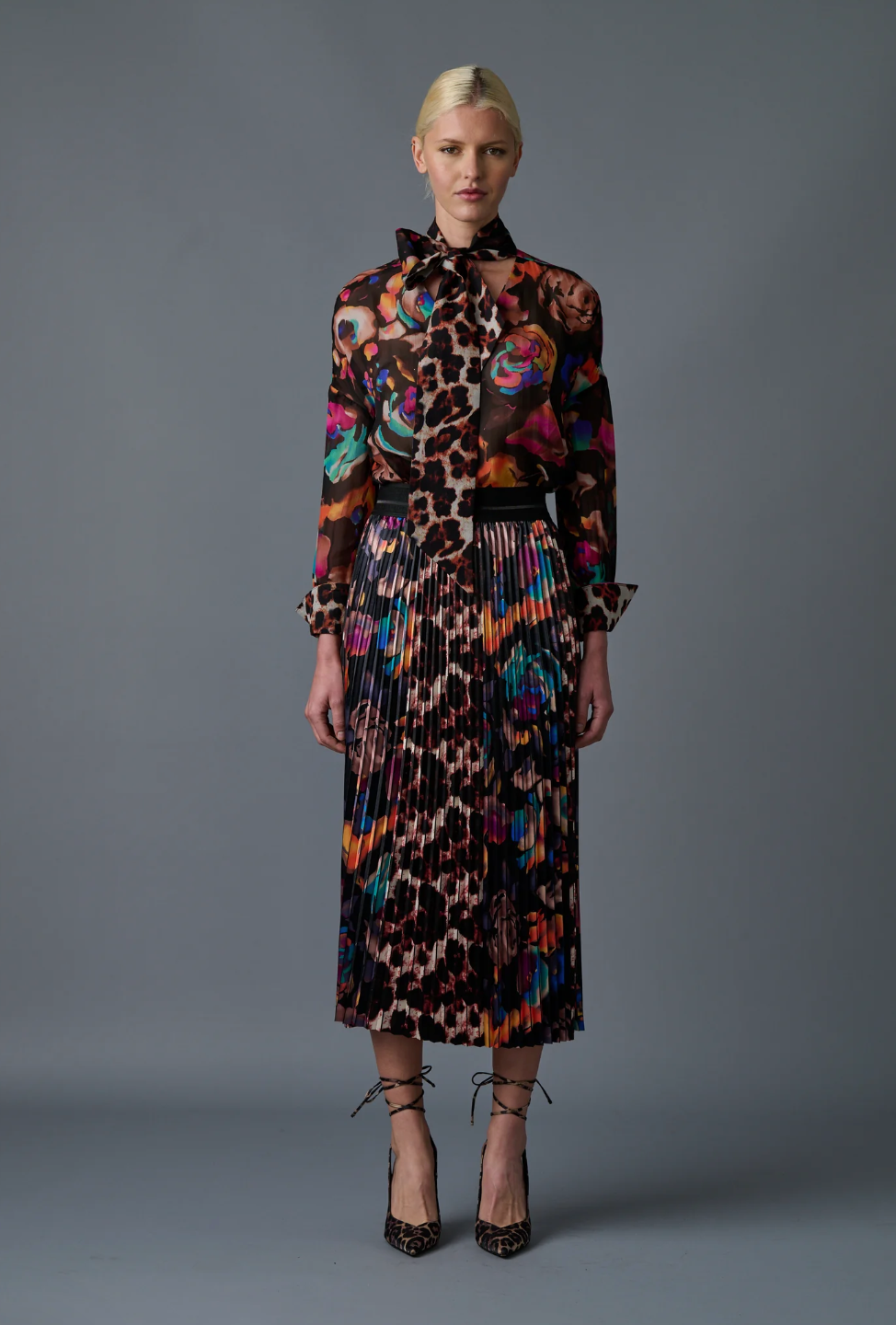 A model with blonde hair tied back is wearing a colorful, long-sleeved, sheer blouse with various abstract patterns paired with the Le Superbe Electric Garden Pleated Skirt featuring an elastic waist and animal print heels. The background is solid gray.