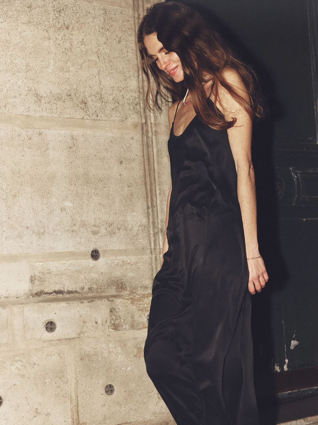 A woman stands against a plain background wearing the Destin Dress by Xirena, a black, sleeveless silk charmeuse dress with adjustable spaghetti straps. She has her arms relaxed behind her and wears black, studded flat shoes. Her long, wavy brown hair cascades over her shoulders as she maintains a neutral expression.