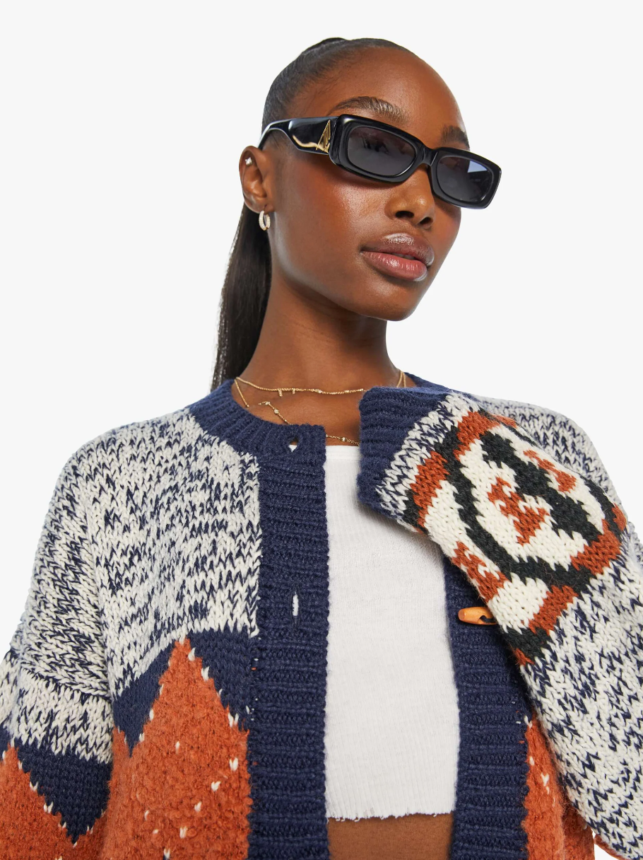 A person with long, pulled-back hair is wearing "The Long Drop Cardigan" by Mother, featuring a patterned design in colors of blue, gray, and orange. They have black sunglasses, a white top, and small hoop earrings. The person is posing with one arm held up, gripping the edge of their cardigan adorned with vintage-inspired wooden buttons.