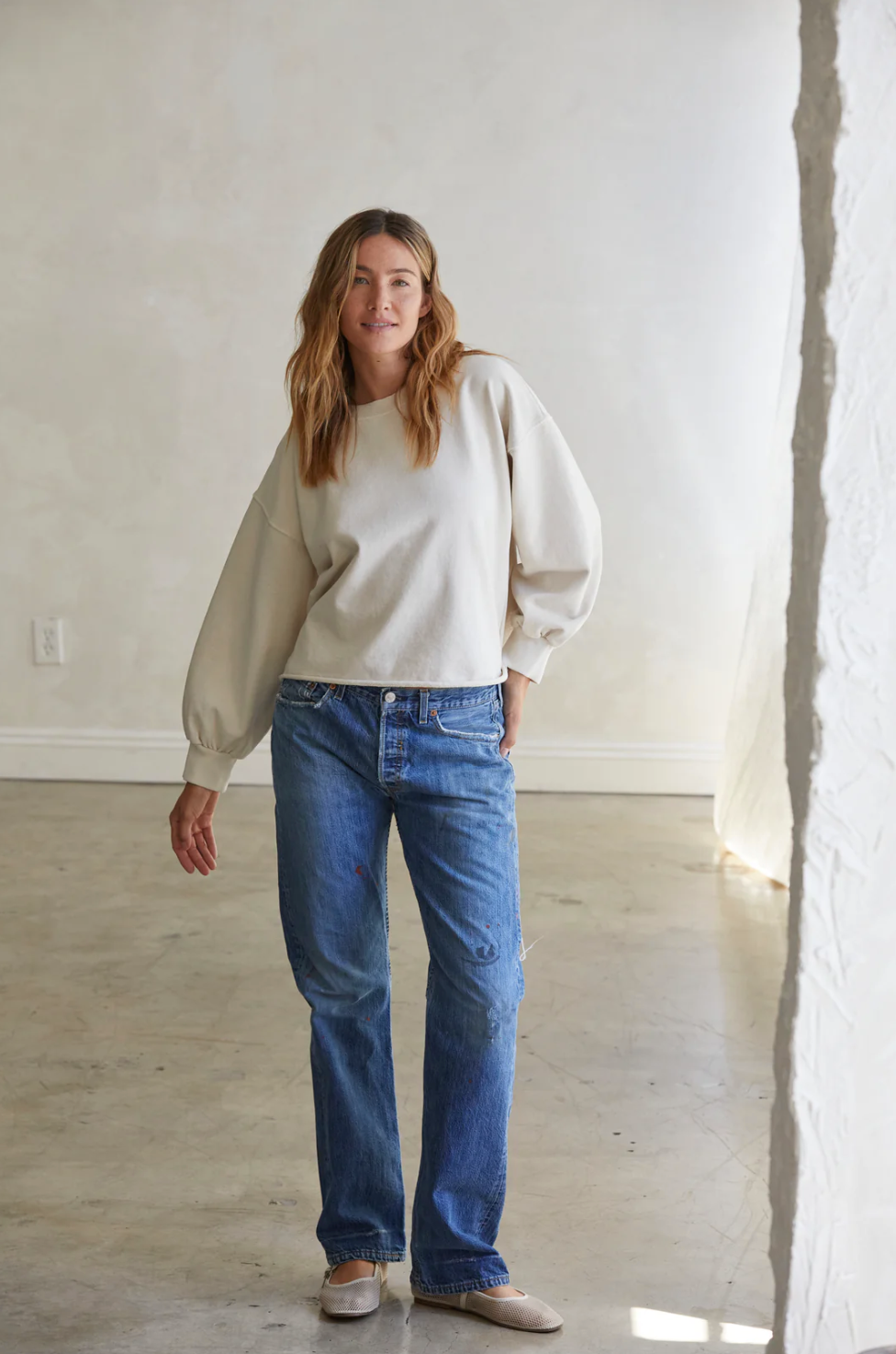 A woman with long wavy hair stands in a minimalistic room with a plain wall and concrete floor. She is wearing a Zayne Terry Pullover by Perfectwhitetee, blue jeans, and beige slip-on shoes. The light-colored french terry fabric of her relaxed silhouette adds to her laid-back look as she gazes at the camera with a relaxed posture, one hand resting at her side.