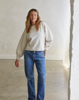 A woman with long wavy hair stands in a minimalistic room with a plain wall and concrete floor. She is wearing a Zayne Terry Pullover by Perfectwhitetee, blue jeans, and beige slip-on shoes. The light-colored french terry fabric of her relaxed silhouette adds to her laid-back look as she gazes at the camera with a relaxed posture, one hand resting at her side.