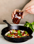 A person is holding a jar labeled "Faire Boon Sauce" and spooning its contents onto a dish in a black cast-iron skillet. The dish appears to have roasted vegetables, cheese, and fresh basil leaves, elevated with added flavor and heat. The background features a bunch of fresh basil on a light-colored surface.