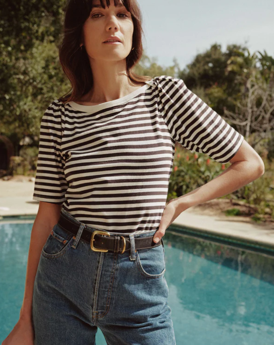 A woman with shoulder-length hair stands by a pool, wearing the Deana Stripe Envelope T-Shirt from Nation LTD. The short-sleeve top is made from organic Peruvian cotton and features black and white stripes. She has tucked it into high-waisted blue jeans, cinched with a black belt that features a gold buckle. Trees and greenery are visible in the background, suggesting an outdoor setting.