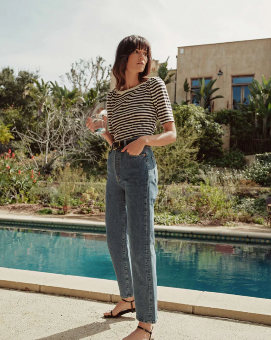 A person with shoulder-length hair stands next to a swimming pool, holding a glass of water. They are wearing the Deana Stripe Envelope T-Shirt by Nation LTD made of organic cotton, high-waisted jeans, and sandals. In the background, there are lush plants, trees, and a large beige building.