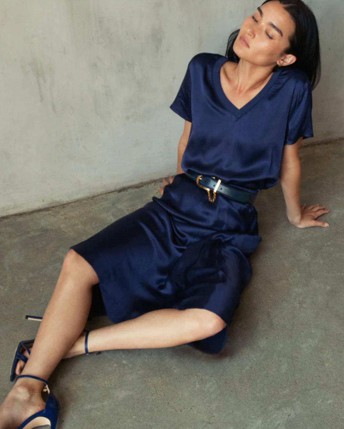 A woman sits on the concrete floor, leaning against a plain wall, wearing a blue V-neck blouse paired with the Mabel Bias Skirt from Nation LTD and a black belt. She has dark hair and blue open-toe heels, with her head tilted slightly back and her eyes closed.