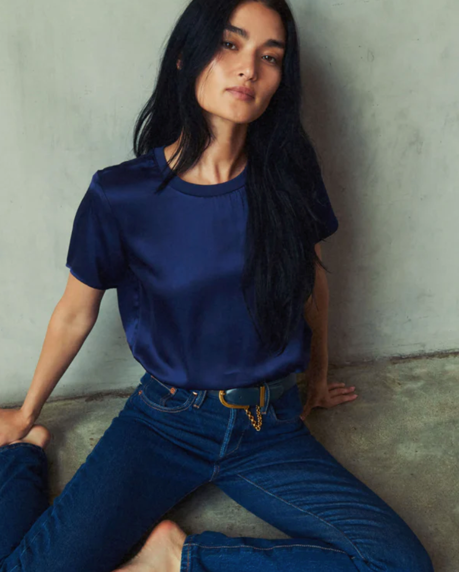 A person with long black hair sits on the floor against a wall, wearing a dark blue Marie Boxy Crop tee by Nation LTD and blue jeans made of lightweight fabric. They are barefoot and looking slightly upward with a calm expression.