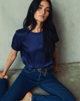 A person with long black hair sits on the floor against a wall, wearing a dark blue Marie Boxy Crop tee by Nation LTD and blue jeans made of lightweight fabric. They are barefoot and looking slightly upward with a calm expression.