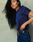 A person with long black hair is standing against a plain light-colored wall, wearing the Marie Boxy Crop, a lightweight navy-blue silk short-sleeve top from Nation LTD, paired with high-waisted blue jeans and a tan leather belt. They are posing with one hand on their hip, looking directly at the camera.