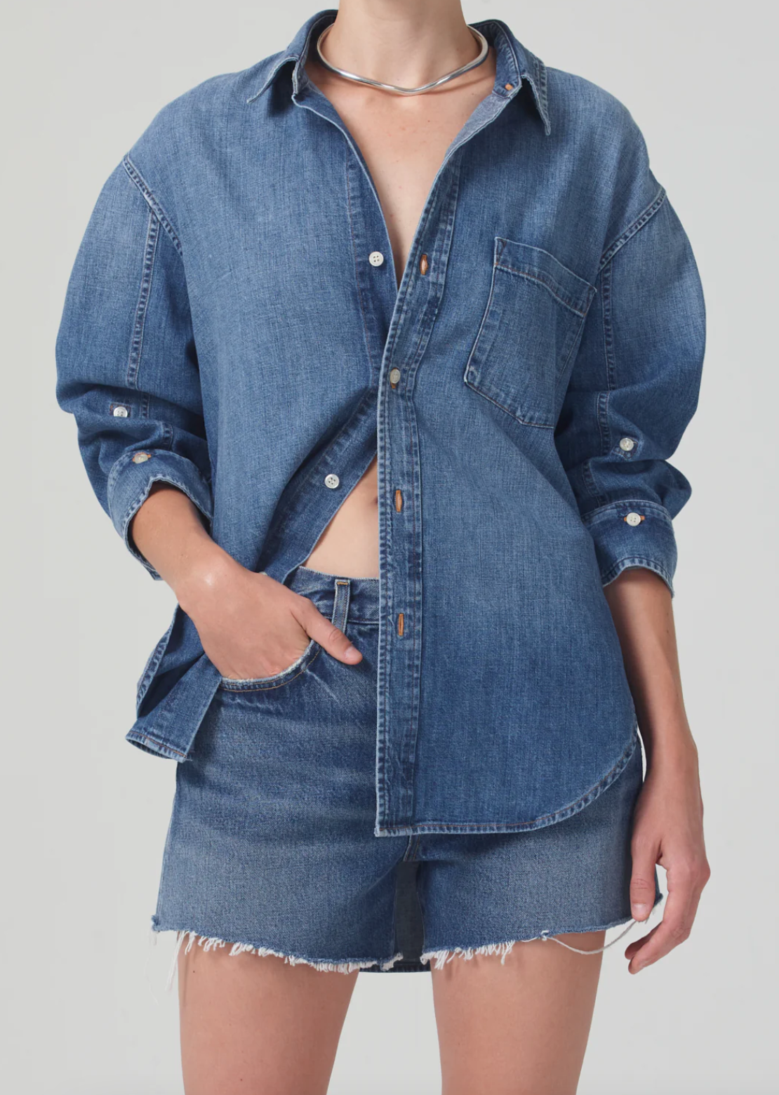 The individual is clad in a Citizens Of Humanity/AGOLDE kayla shirt, which boasts a relaxed fit and long sleeves, adorned with a front pocket. Partially buttoned to show a hint of midriff, the shirt perfectly complements frayed denim shorts. With one hand casually tucked into a pocket, the look is elegantly completed with a silver necklace.