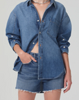 The individual is clad in a Citizens Of Humanity/AGOLDE kayla shirt, which boasts a relaxed fit and long sleeves, adorned with a front pocket. Partially buttoned to show a hint of midriff, the shirt perfectly complements frayed denim shorts. With one hand casually tucked into a pocket, the look is elegantly completed with a silver necklace.