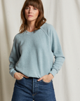 A person with shoulder-length brown hair and bangs stands in front of a light-colored backdrop. They are wearing a cozy essential from Perfectwhitetee, the Ziggy Reverse Fleece LS Crew Sweatshirt in light blue, with the sleeves slightly pushed up. Paired with high-waisted blue jeans, their hands rest comfortably in their pockets.