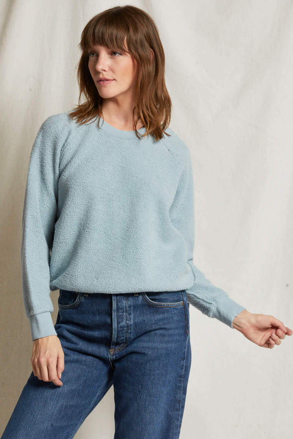 A woman with shoulder-length brown hair is wearing a light blue Ziggy Reverse Fleece LS Crew Sweatshirt by Perfectwhitetee, featuring exposed seam detailing, paired with blue jeans. She is standing against a plain off-white background, looking to her left with one arm bent and her hand slightly raised.