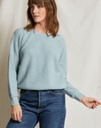 A woman with shoulder-length brown hair is wearing a light blue Ziggy Reverse Fleece LS Crew Sweatshirt by Perfectwhitetee, featuring exposed seam detailing, paired with blue jeans. She is standing against a plain off-white background, looking to her left with one arm bent and her hand slightly raised.