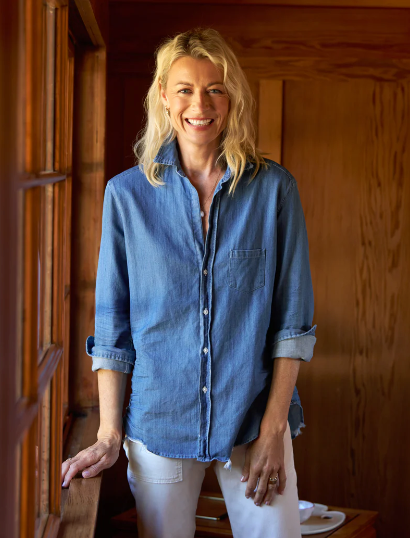 A woman with long blonde hair, wearing a light denim EILEEN Relaxed Button-Up Shirt by Frank & Eileen and light-colored pants, stands by a window in a warmly lit wooden interior. She is smiling and looking at the camera with one hand resting on the windowsill.