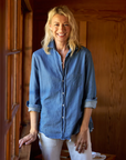 A woman with long blonde hair, wearing a light denim EILEEN Relaxed Button-Up Shirt by Frank & Eileen and light-colored pants, stands by a window in a warmly lit wooden interior. She is smiling and looking at the camera with one hand resting on the windowsill.