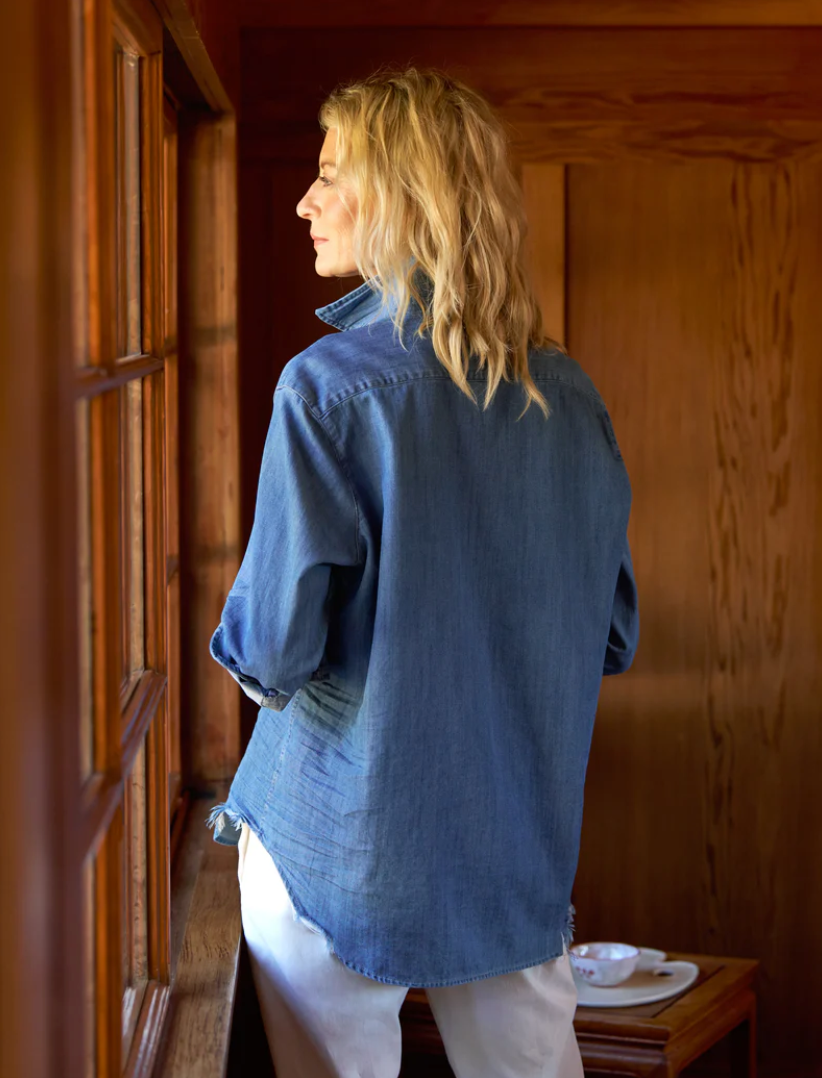 A person with long blonde hair is standing indoors, facing and gazing out of a window. They are wearing the EILEEN Relaxed Button-Up Shirt by Frank & Eileen, known for its bust-flattering button placement, and white pants. The room has wooden walls, and there is a small dish on the wooden surface below the window.