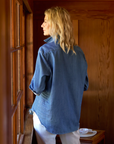 A person with long blonde hair is standing indoors, facing and gazing out of a window. They are wearing the EILEEN Relaxed Button-Up Shirt by Frank & Eileen, known for its bust-flattering button placement, and white pants. The room has wooden walls, and there is a small dish on the wooden surface below the window.