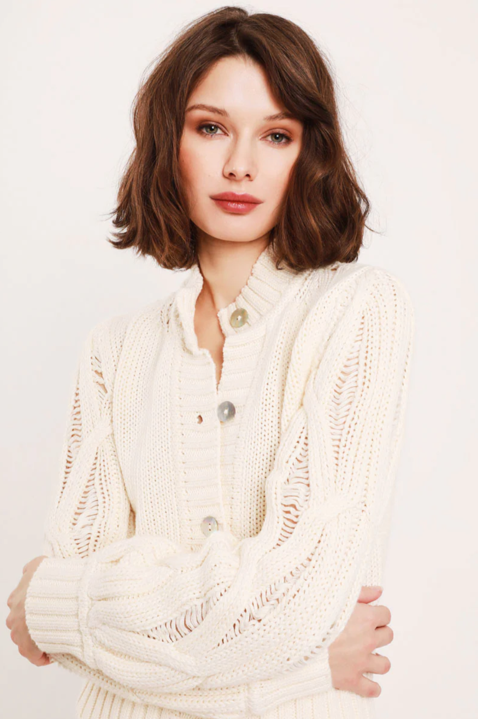 A person with shoulder-length, wavy brown hair wears the Ava Ladder Cable Crop Cardi by Feller, a cream-colored knitted sweater with natural shell buttons. They have a calm expression and are standing in front of a plain, light-colored background.