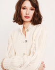 A person with shoulder-length, wavy brown hair wears the Ava Ladder Cable Crop Cardi by Feller, a cream-colored knitted sweater with natural shell buttons. They have a calm expression and are standing in front of a plain, light-colored background.