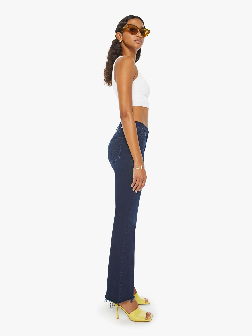 A person with long curly hair is standing sideways, wearing yellow sunglasses, a white sleeveless crop top, The Weekender Fray dark blue wash high-waisted jeans by Mother, and yellow high-heeled shoes. Their right arm is resting at their side.