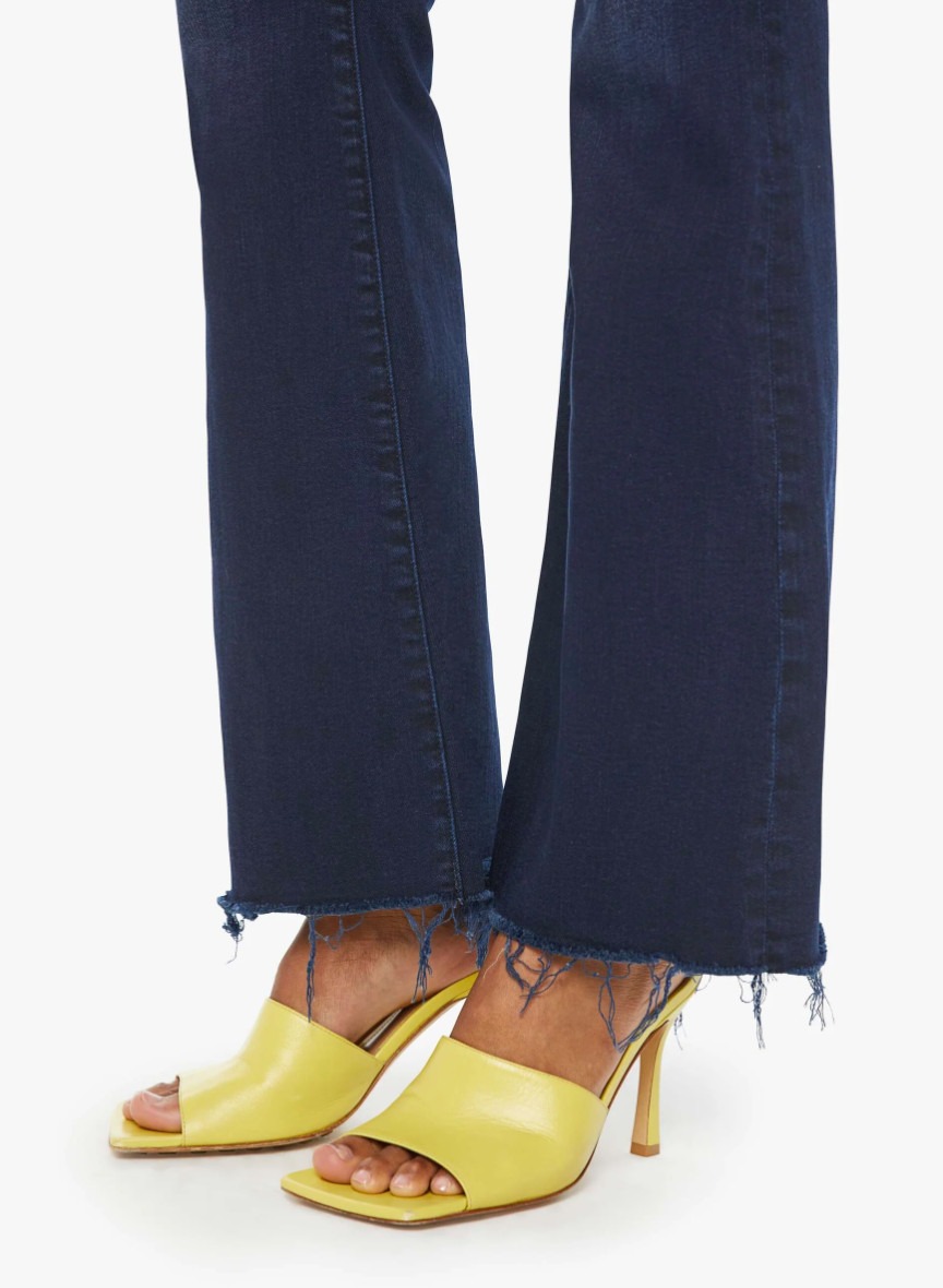 A person wearing The Weekender Fray, a pair of dark blue wash denim jeans by Mother, with a frayed hem and yellow heeled sandals. The focus is on the lower legs and feet, highlighting the stylish combination of mid-rise flare jeans and footwear against a plain white background.