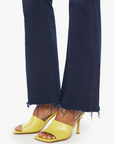 A person wearing The Weekender Fray, a pair of dark blue wash denim jeans by Mother, with a frayed hem and yellow heeled sandals. The focus is on the lower legs and feet, highlighting the stylish combination of mid-rise flare jeans and footwear against a plain white background.