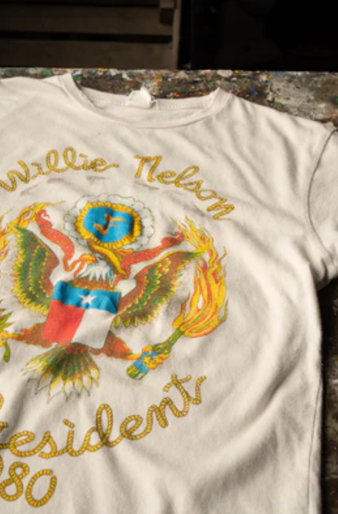 The Willie Nelson For President Tee by Made Worn is laid out on a surface. Crafted from 100% cotton in the USA, this vintage white tee features a vibrant graphic of an eagle holding a flame and sporting a blue shield with a star. The text above the eagle reads "Willie Nelson" and below it reads "President 1980" in eye-catching yellow letters. It's truly iconic.