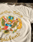 The Willie Nelson For President Tee by Made Worn is laid out on a surface. Crafted from 100% cotton in the USA, this vintage white tee features a vibrant graphic of an eagle holding a flame and sporting a blue shield with a star. The text above the eagle reads "Willie Nelson" and below it reads "President 1980" in eye-catching yellow letters. It's truly iconic.