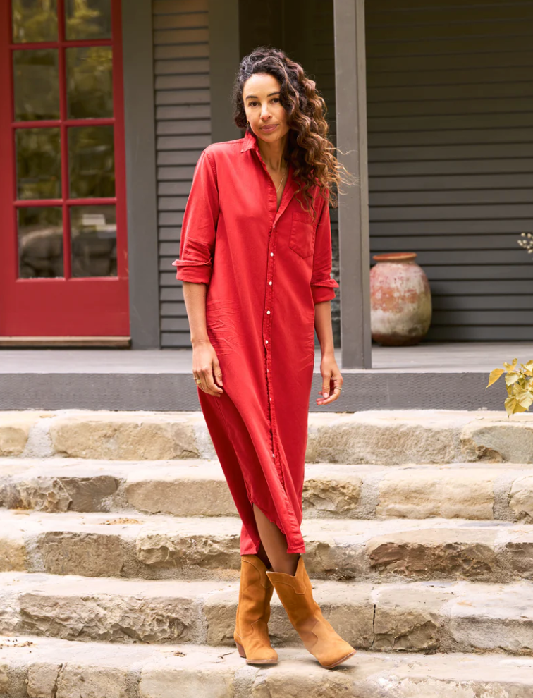 A person with long, wavy hair is outdoors on stone steps, wearing the Rory Shirtdress S24 by Frank & Eileen. They are styled in a red, loose-fit button-up dress paired with brown boots. The backdrop features a building with dark siding and a red door, along with a large ceramic pot visible in the background.