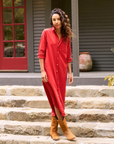 A person with long, wavy hair is outdoors on stone steps, wearing the Rory Shirtdress S24 by Frank & Eileen. They are styled in a red, loose-fit button-up dress paired with brown boots. The backdrop features a building with dark siding and a red door, along with a large ceramic pot visible in the background.