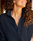 A person with long curly hair is smiling and looking upwards. They are wearing a black Patrick Henley Jersey made of 100% cotton from Frank & Eileen, along with a gold necklace with a pendant. The background is slightly blurred, emphasizing the person's joyful expression and stylish outfit.