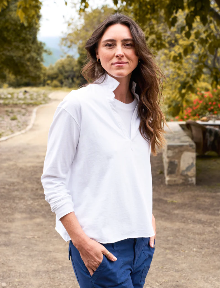 A woman with long, wavy hair stands outdoors on a dirt path, wearing a relaxed fit white Patrick Henley Jersey from Frank & Eileen with the collar turned up and blue pants. The background features trees and foliage, with red flowers visible and a stone bench on the right side of the image.