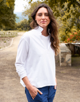 A woman with long, wavy hair stands outdoors on a dirt path, wearing a relaxed fit white Patrick Henley Jersey from Frank & Eileen with the collar turned up and blue pants. The background features trees and foliage, with red flowers visible and a stone bench on the right side of the image.