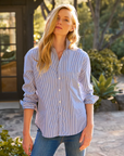 A person with long blonde hair stands outdoors wearing a blue and white striped Eileen Shirt from Frank & Eileen paired with jeans. They have a relaxed expression and stand in a garden with greenery and a building in the background.