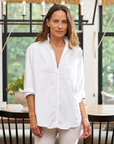 A woman with long brown hair is standing indoors wearing an Eileen Shirt by Frank & Eileen, a white button-down made from 100% linen with rolled-up sleeves. She has a neutral expression and is posed with her arms relaxed at her sides. Behind her is a bright room with large windows, a dining table, and a chandelier.