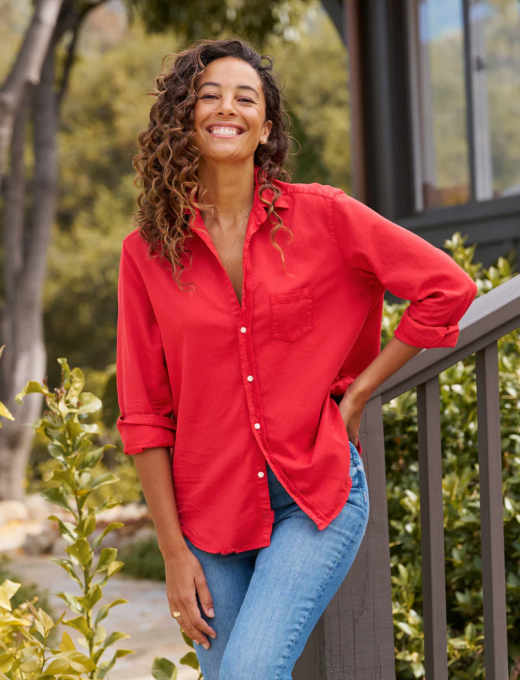 A person with curly hair, wearing an Eileen Shirt by Frank & Eileen and blue jeans, smiles brightly while standing outdoors by a railing. In the background, there are green shrubs and trees, along with the side of a building.