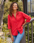 A person with curly hair, wearing an Eileen Shirt by Frank & Eileen and blue jeans, smiles brightly while standing outdoors by a railing. In the background, there are green shrubs and trees, along with the side of a building.