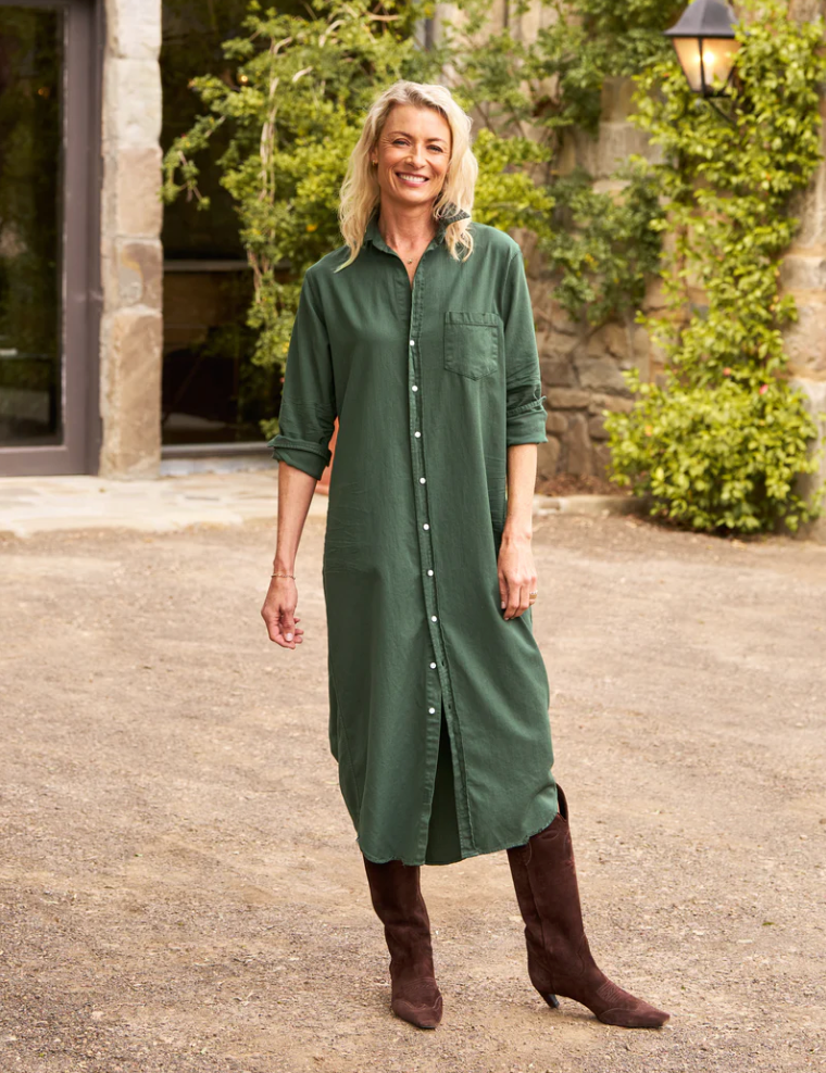 A smiling person stands outdoors in front of a stone building with greenery. They are wearing the Rory Shirtdress S24 from Frank & Eileen, a loose fit, dark green button-up, maxi-length dress paired with dark brown knee-high boots. The linen dress complements their blonde hair as they casually keep one hand in their pocket.