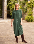 A smiling person stands outdoors in front of a stone building with greenery. They are wearing the Rory Shirtdress S24 from Frank & Eileen, a loose fit, dark green button-up, maxi-length dress paired with dark brown knee-high boots. The linen dress complements their blonde hair as they casually keep one hand in their pocket.