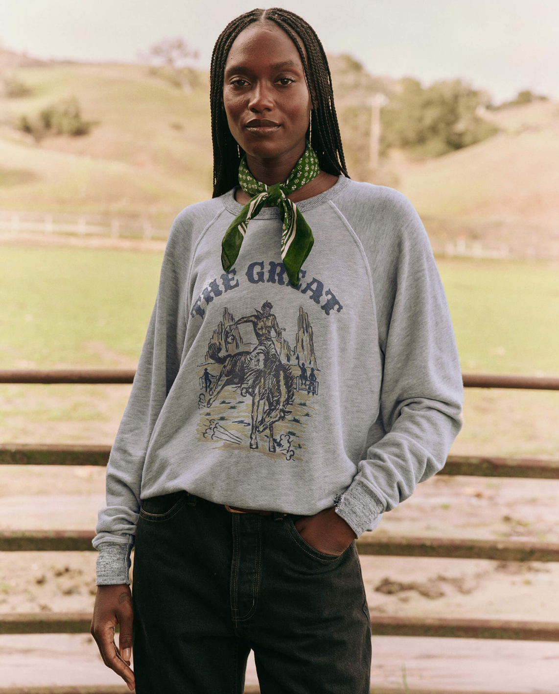 A person with long braids and a green bandana around their neck stands outdoors. They are wearing The Great Inc.&#39;s vintage-style College Sweatshirt, which features an illustration of a cowboy riding a horse and the words &quot;The Great&quot; on it. The background showcases a fenced grassy field and hills, evoking handcrafted in Los Angeles charm.